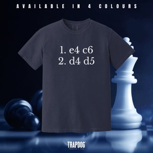 May include: A dark blue t-shirt with white text that reads "1. e4 c6 2. d4 d5". The shirt is photographed against a chessboard background with a white chess piece in the foreground.