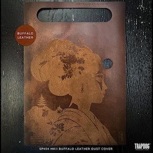 Puede incluir: Una cubierta antipolvo de cuero marrón con un diseño de inspiración japonesa que presenta a una mujer con flores en el pelo. El texto "SP404 MKII BUFFALO LEATHER DUST COVER" está impreso en la parte inferior de la cubierta.