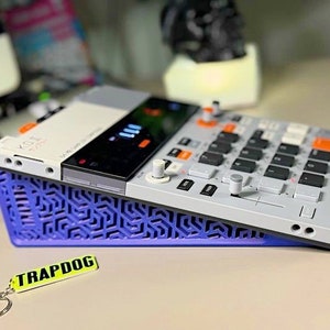 Puede incluir: Una caja de ritmos blanca y gris con botones naranjas y un soporte azul y morado. La caja de ritmos tiene una pequeña pantalla y una variedad de botones y perillas. El soporte tiene un patrón geométrico.