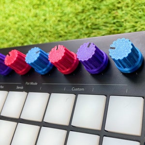 Peut inclure: Un contrôleur de production musicale noir avec six boutons colorés. Les boutons sont étiquetés "Pan", "Sends", "Pot Mode", "Custom".