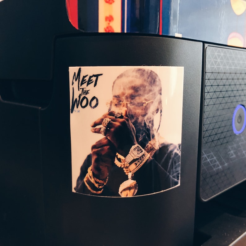 May include: A sticker featuring a portrait of a person with jewelry and the text "Meet the Woo." The person is smoking, with smoke around their face. The sticker is affixed to a black surface.