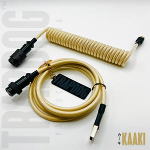 Könnte beinhalten: Gewendeltes beiges Kabel mit schwarzen Steckern und einem USB-C-Anschluss. Das Kabel hat ein schwarzes Etikett mit der Aufschrift TRAPDOOG. Das Wort KAAKI ist auf Japanisch und Englisch geschrieben.