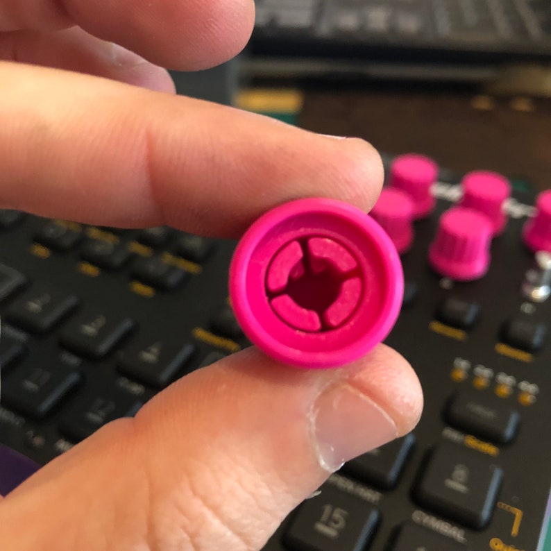 Peut inclure: Gros plan sur un bouton de commande rose vif, avec un design circulaire et une ouverture centrale. Le bouton est tenu entre les doigts, avec d'autres boutons similaires visibles en arri&egrave;re-plan, sugg&eacute;rant un ensemble de composants d'&eacute;quipement audio.