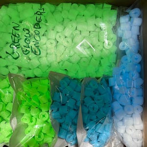 Könnte beinhalten: Nahaufnahme einer Box gefüllt mit Beuteln mit grün, blau und weiß leuchtenden Encoder-Knöpfen. Die Beutel sind mit "Green Glow Encoders" beschriftet.