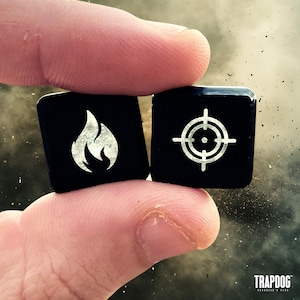 May include: Two black square dice are held between fingers. One die features a silver flame symbol, and the other has a silver target symbol. The dice are from Trapdog.