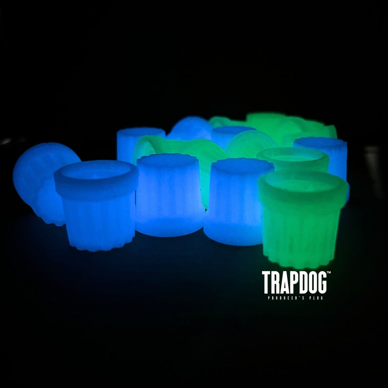 Peut inclure: Un ensemble d'objets cylindriques brillants, bleus et verts, sur fond noir. Les objets ont une surface textur&eacute;e et &eacute;mettent une lumi&egrave;re interne douce. Le texte "TRAPDOG" est visible en bas, avec le slogan "PRODUCER'S PLUGS".
