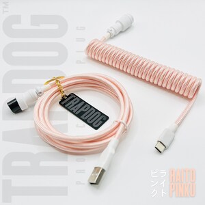 Può includere: Cavo USB rosa a spirale con connettori bianchi e un'etichetta rettangolare nera con la scritta "TRAPDOG". Il cavo è progettato per tastiere meccaniche e altri dispositivi. Il cavo è a spirale e ha un connettore USB-A e USB-C.