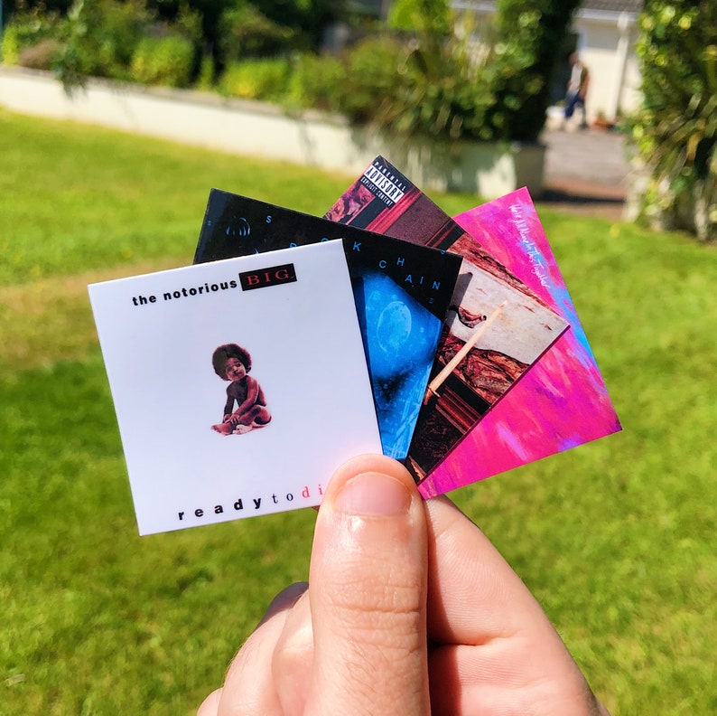May include: A hand holding a fan of album art cards. The top card reads "the notorious BIG" with an image of a baby and the text "ready to die". Other cards feature album art with various colors and designs.