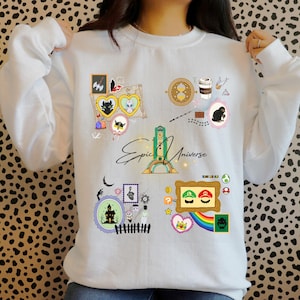 May include: White sweatshirt with colorful graphic designs. The design includes cartoon characters, a coffee cup, a castle, and the words "Epic Universe". The sweatshirt has a crew neck and long sleeves.