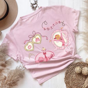 May include: A light pink t-shirt featuring a graphic print with the name LOTTIE in pink circles, a heart-shaped locket containing a frog and a kiss mark, a cartoon character in a pink dress inside an oval frame, and a separate pink ballgown.