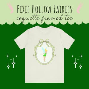 May include: Cream-colored t-shirt featuring a framed illustration of a fairy in a green dress with yellow hair and blue wings. The frame is light green with a bow at the top. The text "PIXIE HOLLOW FAIRIES coquette framed tee" is above the shirt.