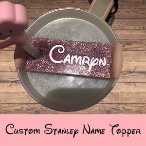 May include: A pink glitter name topper with the name "Camryn" in white lettering. The topper is on a silver Stanley cup lid.