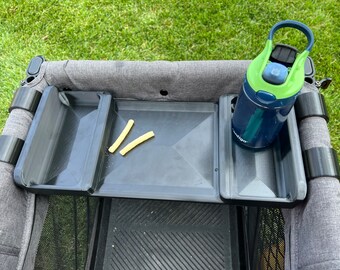 May include: A black stroller tray with two side compartments and a center compartment. The tray has a gray fabric base and a black mesh bottom. A green and blue water bottle is in one of the side compartments. Two yellow sticks are in the center compartment.