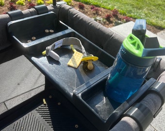 May include: A black stroller tray with a gray fabric handle. The tray has three compartments, one with a pacifier, one with a cracker, and one with a blue water bottle. There are also a few small, round, yellow snacks in the top compartment.