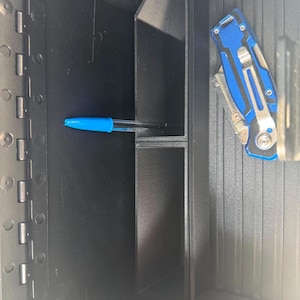 May include: A black storage compartment containing a blue pen and a blue and silver multi-tool. The pen is in a small section, and the multi-tool is in a larger section. The compartment has a textured surface.