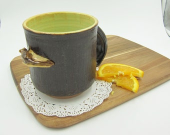 May include: A brown ceramic mug with a unique handle shaped like a bird's beak. The mug has a green glaze on the inside and is sitting on a white doily on a wooden cutting board. There are orange slices on the cutting board.
