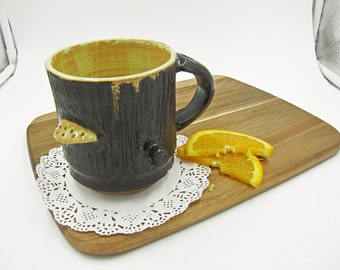 May include: A black and yellow ceramic mug with a tree trunk design and a small bird-shaped handle. The mug is sitting on a white doily on a wooden cutting board with orange slices.