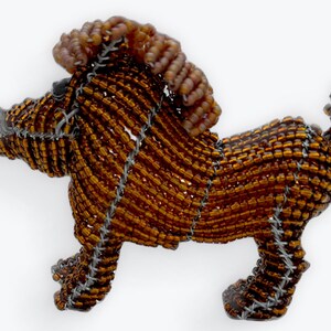 Könnte beinhalten: Eine handgefertigte Löwenfigur aus Perlen. Der Löwe besteht aus kleinen, bernsteinfarbenen Perlen, die sorgfältig mit silberfarbenem Draht aufgefädelt wurden. Die Mähne hat einen helleren Braunton. Die Figur ist ein dekorativer Gegenstand.