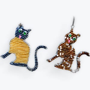 May include: Two beaded cat ornaments. One cat has a yellow and white striped body, blue head and legs, and a black tail. The other cat has a brown and white body, head, and legs, and a black tail. Both cats have green eyes and pink noses.