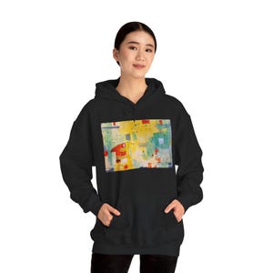 May include: A black hooded sweatshirt featuring a rectangular graphic print on the chest. The print displays an abstract composition with vibrant yellow, red, and teal blue geometric shapes, thin black lines, and small dots on a light background.