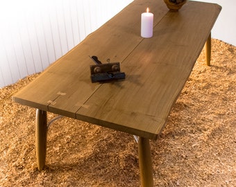 Könnte beinhalten: Ein rustikaler Holztisch mit einer Live-Edge-Platte und konischen Beinen. Der Tisch ist aus hellbraunem Holz gefertigt und hat eine natürliche Oberfläche. Ein kleines Holzhobel und eine weiße Kerze stehen auf dem Tisch.