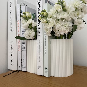 May include: White bookends with a ribbed design holding a bouquet of white flowers. The bookends are holding up a stack of books with titles like "Urban Botanics", "Home", and "The New Plant Parent".