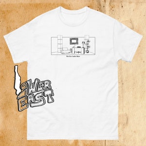 Puede incluir: Camiseta blanca con una ilustración en blanco y negro de un plató de estudio de televisión. El texto "The Eric Andre Show" está impreso debajo de la ilustración.