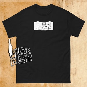 Puede incluir: Camiseta negra con un gráfico blanco de una habitación con muebles y plantas. El texto "The Eric Andre Show" está debajo del gráfico. El texto "Lower East" está en la parte inferior izquierda de la camiseta.