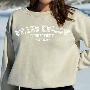 May include: A light beige sweatshirt with a white graphic print that reads "Stars Hollow Connecticut - Est. 1779 - "