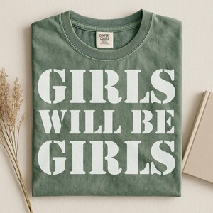 Peut inclure: T-shirt vert sauge plié avec l'inscription blanche « GIRLS WILL BE GIRLS ». Le t-shirt est posé sur une surface neutre, avec une branche de fleurs séchées et un livre en arrière-plan. Col rond, manches courtes.