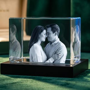 May include: A rectangular glass block featuring a laser-etched 3D monochrome portrait of a couple embracing. The glass block is mounted on a solid, dark rectangular base, creating a clear display of the detailed photographic engraving inside.