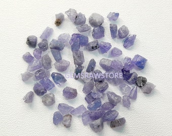 May include: A collection of raw tanzanite gemstones in various shapes and sizes. The stones display shades of blue and purple, with a rough, natural texture. The image is taken on a white surface, with the text "GEMSRAWSTORE" visible.