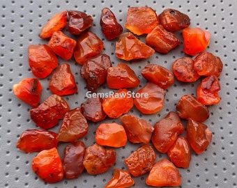 May include: A collection of approximately 30 carnelian gemstone roughs. The carnelian stones are a mix of orange and red hues. The stones are irregularly shaped and have a natural, unpolished finish.