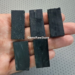 May include: Five rectangular, raw black tourmaline stones. The stones have a rough, textured surface and varying sizes. The image is taken from a close-up angle, showcasing the natural beauty of the crystals. The stones are held in a hand.