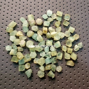 May include: A collection of small, light green aquamarine crystals. The crystals are rough and have a natural, unpolished look. They are scattered on a textured surface.