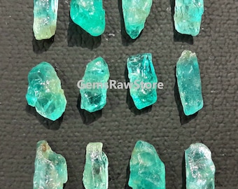 Può includere: Una collezione di dodici gemme di acquamarina grezza. Le pietre, di varie dimensioni e forme, mostrano sfumature di verde-blu e turchese. Le gemme sono disposte su una superficie scura e strutturata. Il testo "GemsRawStore" è nell'immagine.