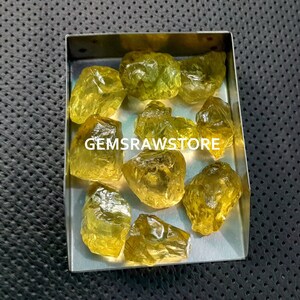 May include: A collection of yellow, raw, uncut gemstones in a metal tray. The gemstones are various sizes and shapes. The text "GEMSRawSTORE" is visible in the image.