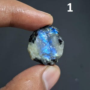 May include: A raw, iridescent labradorite gemstone with flashes of blue and black. The rough stone is held between fingers, showcasing its natural, unpolished texture. The number "1" is in the upper right corner.