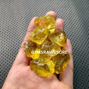 May include: A collection of yellow citrine crystals, raw and unpolished, in a hand. The crystals are various sizes and shapes, with some having a more translucent appearance than others.  GEMSRawSTORE is printed on the image.