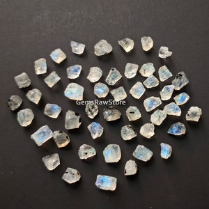 Puede incluir: Una colección de cristales de piedra lunar en bruto, con colores iridiscentes azules y blancos. Los cristales son de varias formas y tamaños, y algunos tienen inclusiones negras.