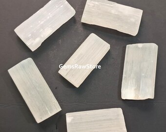 May include: Six rectangular selenite crystal wands, pale translucent white, arranged on a dark surface. The wands have visible striations and a natural, raw appearance. The text "GemsRawStore" is visible on the surface.