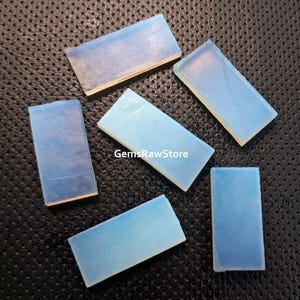 May include: Six rectangular pieces of opalite, a translucent, milky white material, with a slight iridescent sheen. The pieces are arranged on a black textured surface.  GemsRawStore is printed on one of the pieces.
