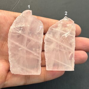 Könnte beinhalten: Zwei durchscheinende, blassrosa Rosenquarz-Kristallscheiben mit unregelmäßigen, gezackten Kanten und sichtbaren inneren Brüchen. Das linke Stück ist mit der Nummer 1 und das rechte Stück mit der Nummer 2 gekennzeichnet, beide mit einer glatten Oberfläche.