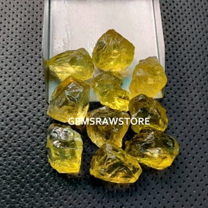 May include: A group of yellow citrine rough gemstones in various sizes and shapes. The gemstones are displayed on a white tray with a black background.