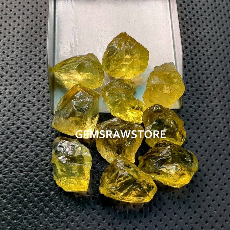 May include: A group of yellow citrine rough gemstones in various sizes and shapes. The gemstones are displayed on a white tray with a black background.