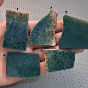 May include: Five irregularly shaped, polished stone slabs in shades of teal and green are displayed. Each piece has a unique shape and colour variation, with some showing brown and beige edges. The stones are held in a hand against a neutral background.