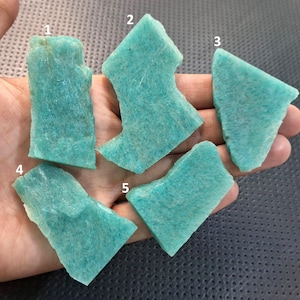 Könnte beinhalten: Fünf Stücke rohen Amazonit-Edelsteins in verschiedenen Formen und Größen. Der Amazonit hat eine helltürkise Farbe mit einer leicht rauen Textur. Die Edelsteine werden in einer Hand vor einem dunklen Hintergrund gehalten.
