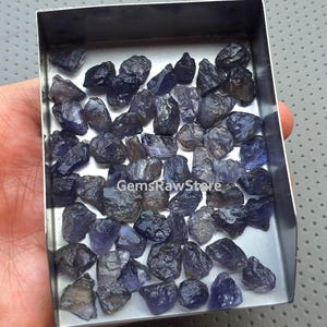 Könnte beinhalten: Eine Schale gefüllt mit zahlreichen grob geschnittenen, dunkelblau-violetten Iolith-Edelsteinen. Die rohen Steine variieren in Größe und Form und weisen eine natürliche, unpolierte Textur auf. Das Bild ist gut beleuchtet und hebt die Farbe und Kristallstruktur des Ioliths hervor.