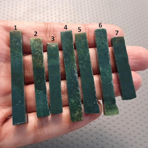 Puede incluir: Siete piezas rectangulares de piedra verde oscuro de diferentes longitudes se sostienen en una mano. Las piedras tienen un aspecto pulido y algunas tienen motas verde claro y amarillo-marrón. Las piedras están dispuestas verticalmente.
