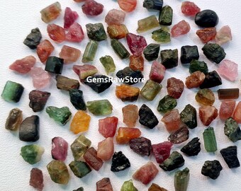 Könnte beinhalten: Eine Sammlung roher, unpolierter Turmalin-Edelsteine in verschiedenen Grüntönen, Rosa, Schwarz und Rot. Die Edelsteine sind roh und haben eine natürliche, unregelmäßige Form.